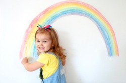 Super cute little girl drawing rainbow on white wall at home. Sign of hope. Quarantine. Coronavirus. Smiling child looking in camera. Stay at home.