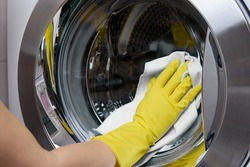 In the bathroom, a cleaner wipes a steel-colored washing machine drum with a white rag. A woman's hand in a yellow latex glove wraps the drum of a washing machine to a shine with a green rag