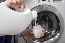 Against background of drum of steel-colored washing machine, woman pours liquid washing gel into plastic cap. A girl in a white T-shirt carefully pours a transparent conditioner for flattening laundry