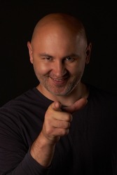 Laughing bald man on a black background points his finger at the camera, vertical photo. A smiling, sly man points his index finger at the camera. You are being deceived.
