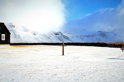wooden cross in the cemetery, stone fence of the black church in Budir and in the background snow-capped and misty mountains, Iceland