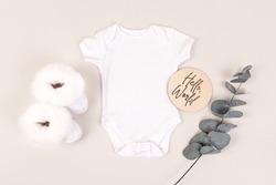 White baby bodysuit with white furry baby shoes and green eucalyptus leaf. Hello world wooden baby milestone sign. Minimal and modern white baby onesie mockup.