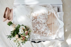 Luxury wedding dress in white box, beige women's shoes and bridal bouquet on bed, copy space. Bridal morning preparations. Wedding concept