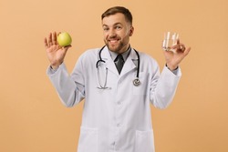 The male nutritionist doctor with stethoscope smiling and holding water and apple on beige background, diet plan concept
