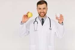 The happy male nutritionist doctor with stethoscope smiling and holding water and apple on white background, diet plan concept