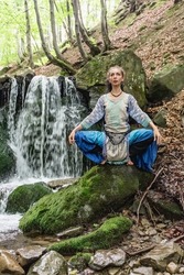 brunette woman in blue clothes sits near a mountain waterfall, Carpathian summer forest, Ukraine

