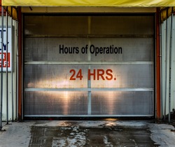 Hours of operation (24 hours) vinyl letter and number signage on a car wash with headlights shining through roll door. Wet pavement in foreground.