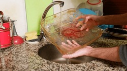 At the kitchen sink, the man lathers a dirty glass casserole dish and washes the dishes by hand.