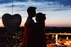 Boy and girl hug each other tender standing on the rooftop in the rays of evening lights and having a romantic dinner
