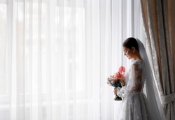Side view of fashionable bride, woman with long veil wearing in wedding dress with long sleeves decorated with lace, and looking at bouquet of flowers while standing in front window indoors