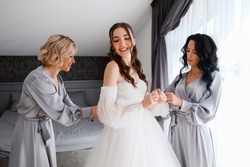 Crop portrait of pretty elegant woman bride in puffy white dress smiling while bridesmaids helping prepare for wedding ceremony, standing spacious room in silk gray robe. Morning of bride.