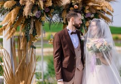 Front view of wedding ceremony of groom in brown tuxedo and bride with wedding veil on face wearing in puffy dress, looking at each other while standing near decorated arch with dried flowers