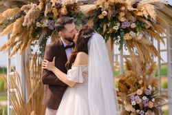 Cropped of loving brides couple, elegant bride with veil, wearing in wedding dress kissing with stylish groom in brown suit while standing together near lake and decorated with dried flowers gazebo