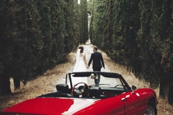 Front view of red oldfashioned convertible car on foreground and back view of walking on narrow path bride and groom holding together hands. beautiful couple in love. Concept of love and marriage.