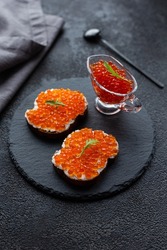 Red caviar in a bowl and caviar sandwiches on a black stone board. On a black structural background