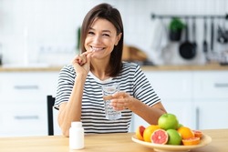 Young beautiful smiling caucasian woman holding vitamin pill and glass of water, looking at the camera and smiles friendly. Healthy lifestyle, healthy diet nutrition concep