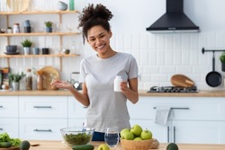 Happy african american woman in home kitchen holding a bottle of nutritional supplements, looking at the camera and smiling friendly, healthy lifestyle