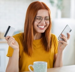 Young cheerful caucasian woman using bank credit card and mobile phone to shopping online on the internet, redhead beautiful excited girl feels euphoria from long-awaited purchase the Internet