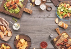 Outdoors Food Concept. On the wooden table different food with copy space, grilled chicken legs, buffalo wings, bread, salad, potatoes, potato chips and beer