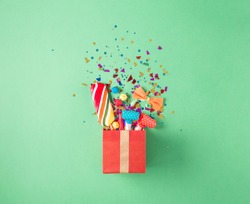 Red gift box with various party confetti, balloons, streamers, noisemakers and decoration on a green background. Flat lay