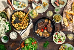 Dinner table with meat grill, roast new potatoes, vegetables, salads, sauces, snacks and lemonade, top view