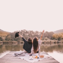 Two female friends in knitted warm sweaters having picnic near lake with autumn forest and lake on the background. Cozy fall atmosphere. Place for text.