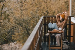 Young woman in knitted sweater and hat drinking tea and eating fresh croissants on cozy balcony of a wooden country house on autumn day. 