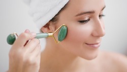 Beautiful young woman with perfect skin wearing towel on head using a jade face roller with natural quartz stones on white background.