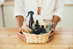 Woman holding wicker basket with natural eco-friendly zero waste plastic free cleaning items: brushes, rugs, soap, essential oils, spray and sponges at the kitchen. Plastic free home.