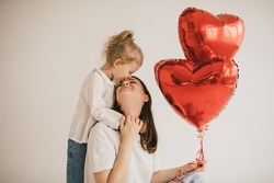 Young mother and her little daughter holding red heart shaped balloons on white background. Saint Valentine's Day celebration.
