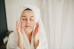 Young beautiful woman wearing white bathrobe and towel on her hair applying moistrizing cream on her face. Skin care morning rituals. Beauty routine. 
