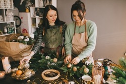 Two millennial women making Christmas wreath using fresh pine branches and festive decorations. Small business