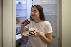 Woman smiling enjoys a delicious sweet chocolate cake at night alone near the fridge. Funny photo. A woman with trouble eating. Carbohydrates, calories. Nocturnal Harmful Forbidden Food