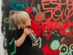 Caucasian blonde toddler looking at the graffiti he has just painted on the wall with a red spray