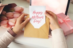 Woman's hands holding a letter in craft envelope. Pink background, Mother's day concept. Tulips flower and pink gift box in background. Womens home desk.