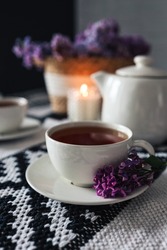 A cup of tea with a branch of lilac on a saucer on the table. Candle, teapot, wicker basket with lilacs. Aesthetics of tea drinking.