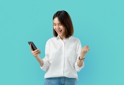 Young smiling Asian woman holding smart phone with fist hand and excited for success on light blue background.