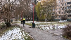 In the city park, wet snow on the grass, a shop courier rides a bicycle along the wet path of the park with a travel backpack on his bac