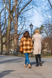 Two girls walk along an asphalt path past tall trees in the city in early spring