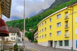 The road past the colorful houses on a rainy May evening on the street of Liechtenstein's capital Vaduz against the backdrop of a green mountain forest and a cloudy sky over the Alps