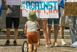 A little boy on a balance bike against the background of adult activists of the anti-war protest of free Russians against military aggression on a sunny day on a cobbled old square in Krakow, Poland