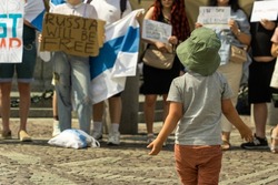 A little boy in panama stands in front of placards in the hands of adult activists of the anti-war protest of free Russians against military aggression on a sunny day in the cobbled old square