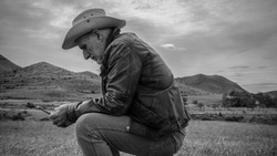 Monochrome of adult man in cowboy hat sitting on field using cellphone
