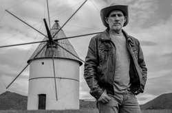 Portrait of adult man in cowboy hat standing on field with Spanish traditional windmill against sky
