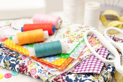 sewing, sewing on the sewing machine, sewing supplies, colored sewing threads, colored pieces of cloth, needles, centimeter, tailors scissors on white wooden background