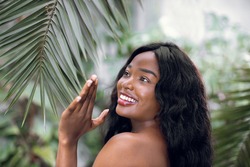 Concept of natural beauty and skincare. Young pretty African woman with perfect smooth skin and long hair, standing in beautiful greenhouse and touching leaf of palm tree.