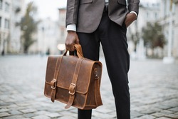 Close up of successful african businessman in suit standing on city street with brown leather briefcase in hands. Young busy worker walking outdoors.