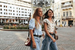 Blonde and brunette women in stylish loose jeans and floral trendy blouses walk and talk in good mood outside. Portrait of attractive girlfriends.