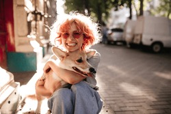 Happy curly woman in jeans smiles sincerely and hugs corgi. Good-humored girl in red sunglasses plays with dog on street.