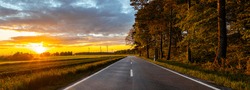 A panoramic shot of a country road on the side of an agricultural field against a beautiful sunset sky